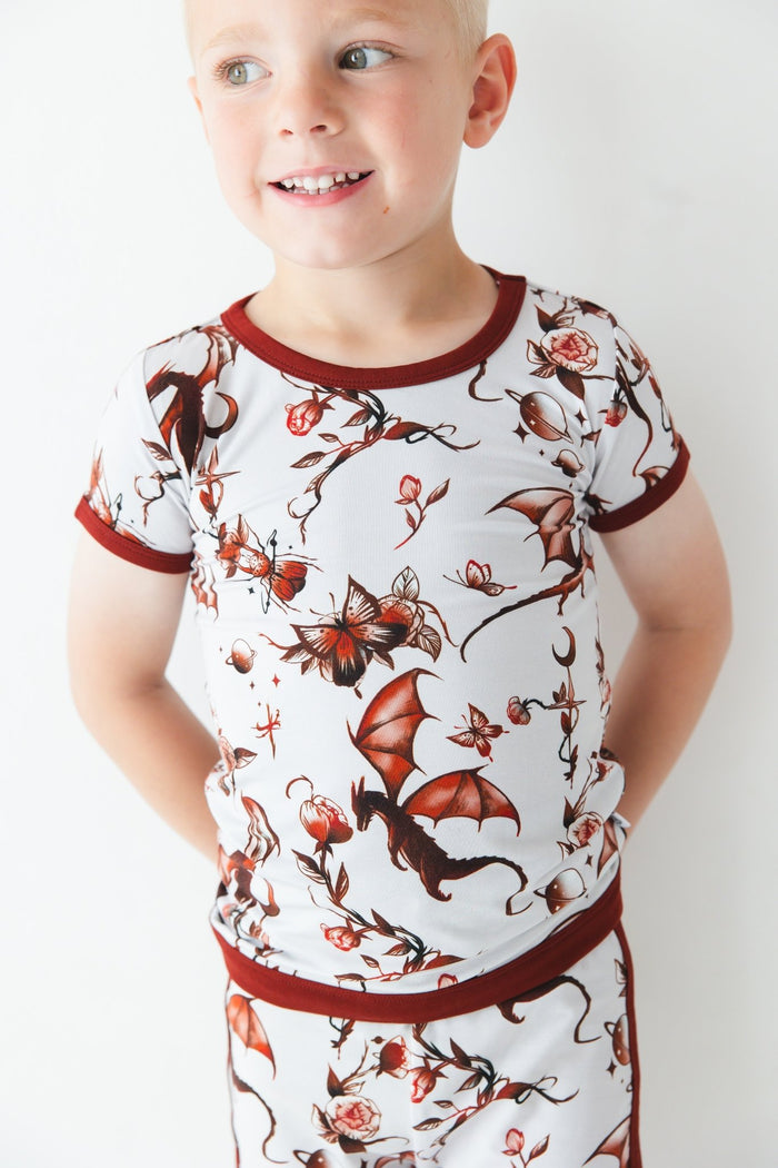 A young child wears the Jammie Session Dreaming with Dragons Short Pajama Set‚Äîbreathable kids‚Äô pajamas featuring a matching short-sleeve top and shorts with a brown dragon and floral print‚Äîposed against a white background.