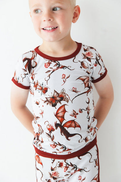 A young child wears the Jammie Session Dreaming with Dragons Short Pajama Set‚Äîbreathable kids‚Äô pajamas featuring a matching short-sleeve top and shorts with a brown dragon and floral print‚Äîposed against a white background.
