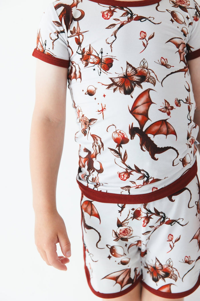 A child in the Jammie Session Dreaming with Dragons Short Pajama Set‚Äîfeaturing a matching short-sleeve shirt and shorts with red dragon and floral prints‚Äîstands against a plain white background.