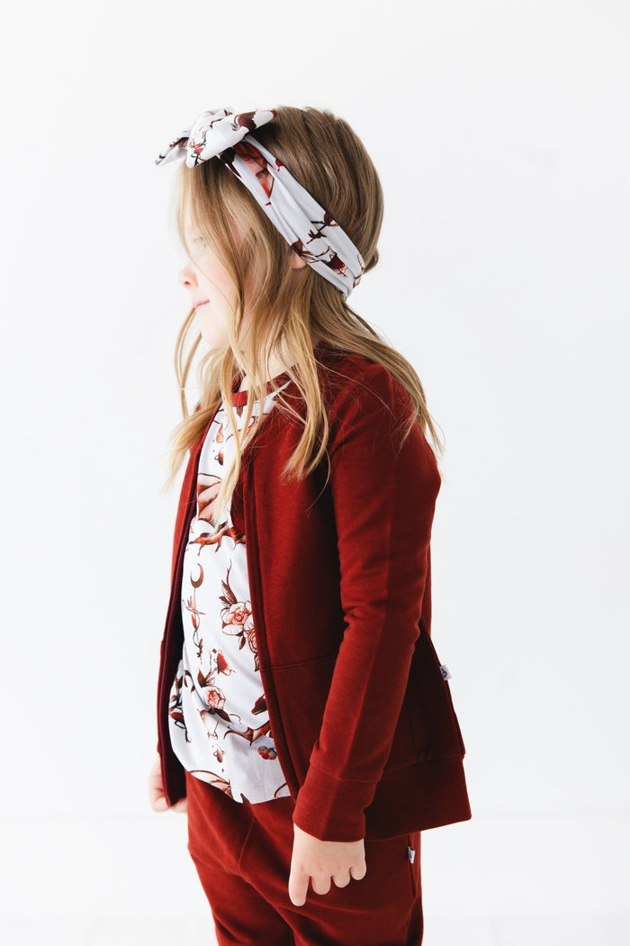 A young child with long hair wears the Jammie Session Dreaming with Dragons Solid Kids' Jacket (runs small, please size up) in red and matching pants, paired with a floral shirt and headband, standing against a plain white background.