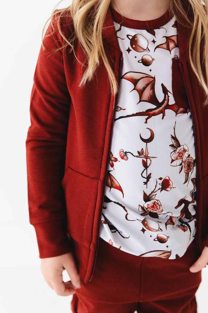 A child in a maroon Dreaming with Dragons Solid Kids' Jacket by Jammie Session, paired with pants and a white dragon and planet print shirt.