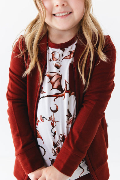 A young child wears the Jammie Session "Dreaming with Dragons" Solid Kids' Jacket (runs small, please size up) in dark red over a white shirt with flying dragons, moons, and stars, standing against a plain light background.