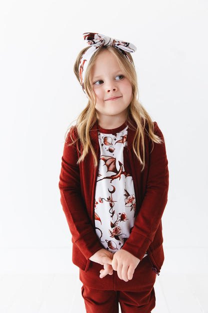 A young girl stands against a white background, smiling gently and wearing the Jammie Session Dreaming with Dragons Solid Kids' Jacket (runs small, please size up) paired with a floral headband.