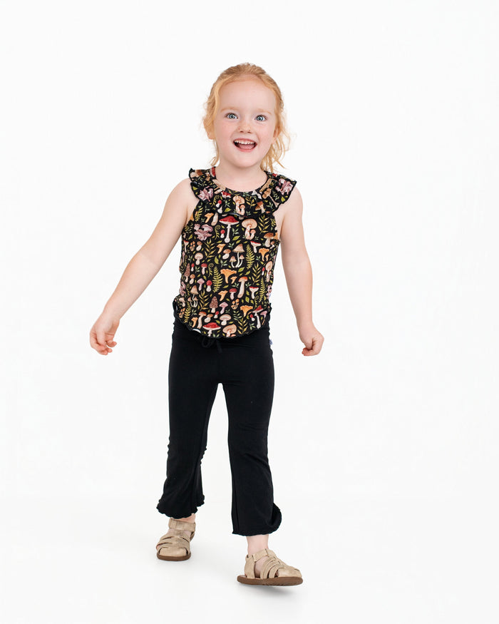 A young child with light hair smiles while wearing the Jammie Session Mystic Mushrooms Bloomy Crop Set, featuring a sleeveless patterned top and black pants, standing against a plain white background.
