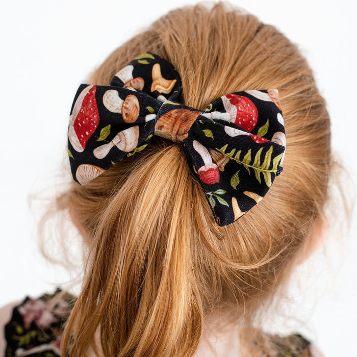 A blonde-haired girl seen from behind wears the Jammie Session Mystic Mushrooms Bow, a black hair bow featuring a charming mushroom and foliage pattern.