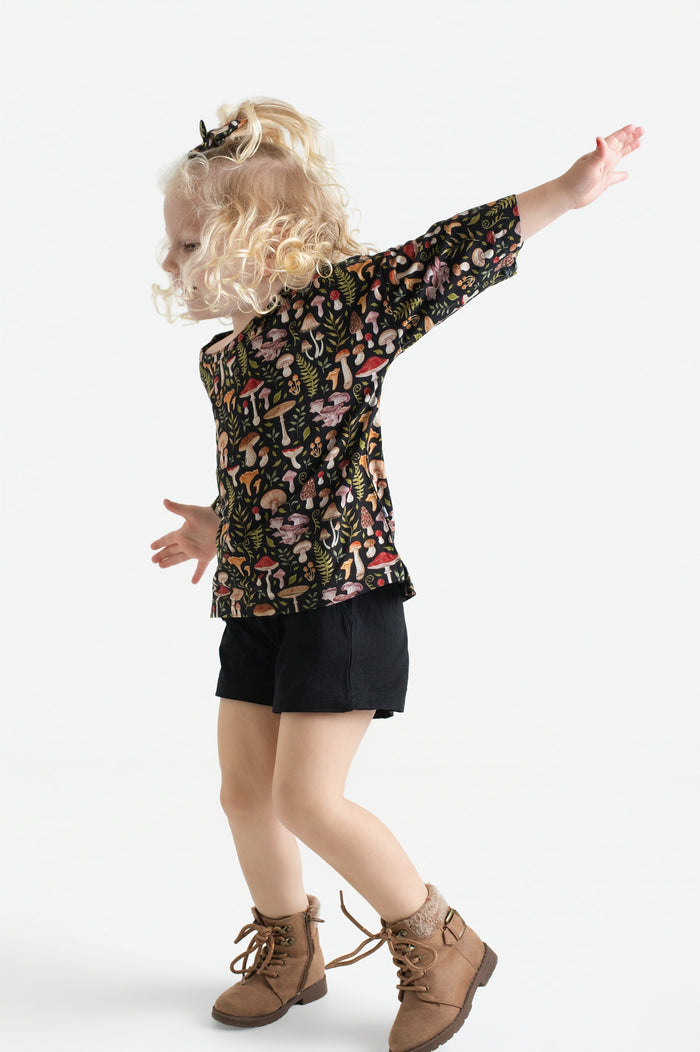 A young child with curly blonde hair models the Jammie Session Mystic Mushrooms Comfy Cloud Short Set, standing with arms outstretched against a plain white background.