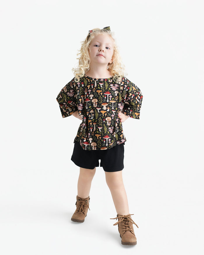 A young child poses confidently in the Jammie Session Mystic Mushrooms Comfy Cloud Short Set, featuring a patterned top and black shorts, paired with brown boots and a matching hair accessory against a plain white background.