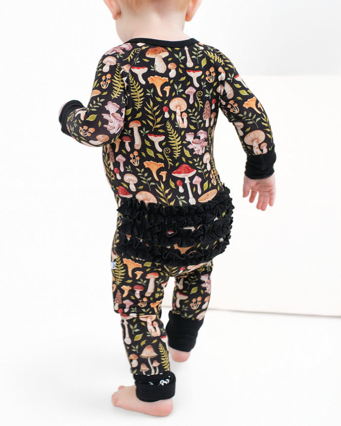 A toddler in Jammie Session's Mystic Mushrooms Ruffle Romper, a black bamboo outfit with a vibrant mushroom and fern pattern plus ruffled back, walks barefoot on a white surface.