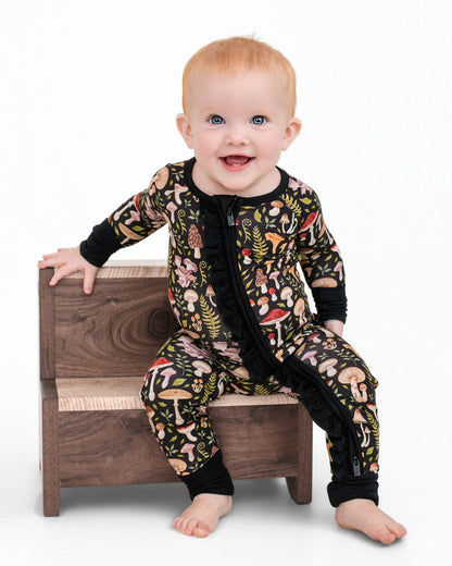 A baby with light hair sits on wooden steps, wearing the Jammie Session Mystic Mushrooms Ruffle Romper, which has a black base and a colorful mushroom forest print, set against a plain white background.
