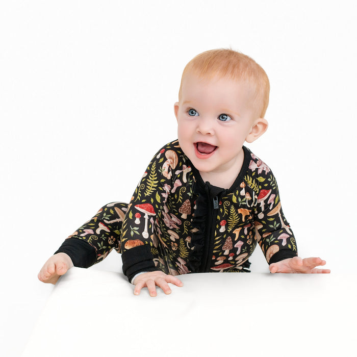 A baby with light hair crawls on a white surface, dressed in the Jammie Session Mystic Mushrooms Ruffle Romper‚Äîa black long sleeve romper featuring colorful mushroom and plant prints‚Äîagainst a plain white background.