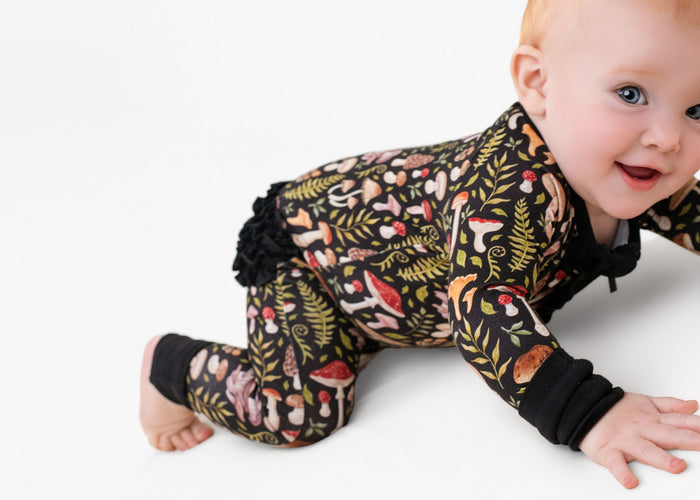 A baby crawls on a white surface in the adorable Mystic Mushrooms Ruffle Romper by Jammie Session, featuring a long-sleeve design with a colorful mushroom and foliage pattern.