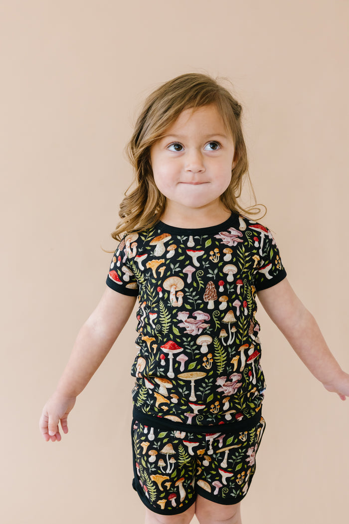 A young child in Jammie Session‚Äôs Mystic Mushrooms Short Pajama Set stands against a beige background‚Äîthese breathable bamboo pajamas feature a colorful mushroom pattern and are gentle on sensitive skin.