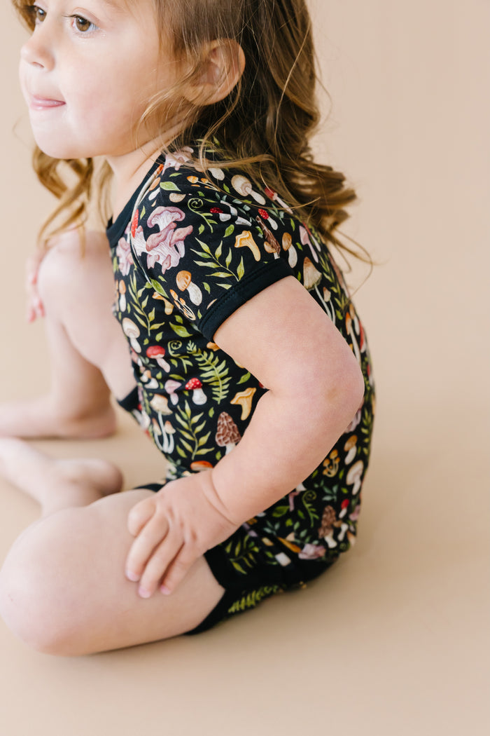 A young child with brown hair sits on a beige surface, wearing the Jammie Session Mystic Mushrooms Short Pajama Set‚Äîbreathable kids‚Äô sleepwear with a vibrant nature-inspired print for ultimate comfort.