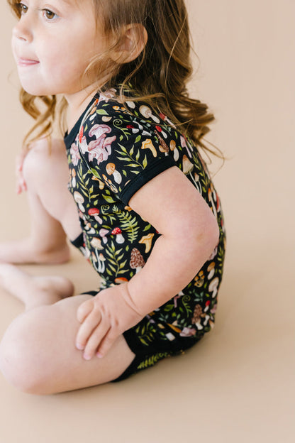 A young child with brown hair sits on a beige surface, wearing the Jammie Session Mystic Mushrooms Short Pajama Set‚Äîbreathable kids‚Äô sleepwear with a vibrant nature-inspired print for ultimate comfort.