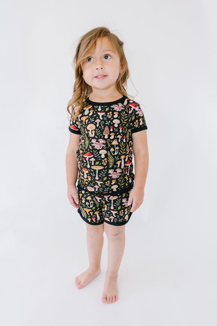 A young child stands against a white background, wearing the Jammie Session Mystic Mushrooms Short Pajama Set‚Äîsoft, breathable kids pajamas in black with a vibrant mushroom print for all-day comfort.