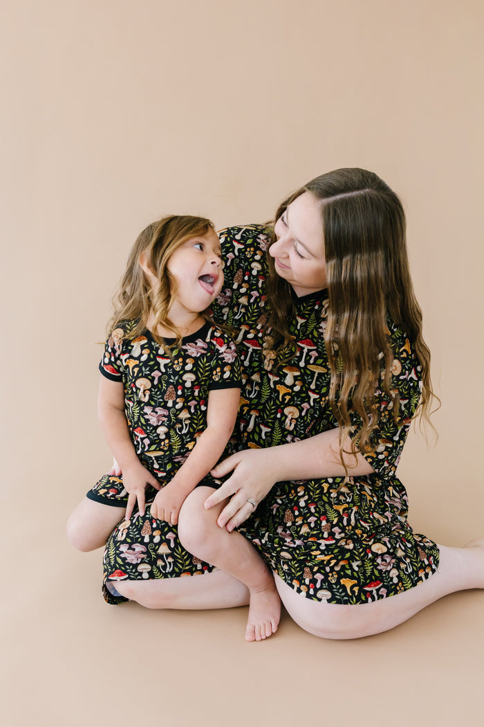 A woman and a young girl sit on the floor against a beige background, smiling at each other in matching Jammie Session Mystic Mushrooms Short Pajama Sets‚Äîstylish, hypoallergenic kids' clothing with colorful patterns.