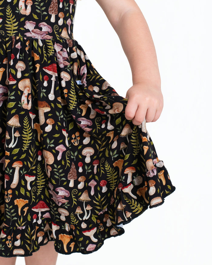 A child holds out the skirt of the Jammie Session Mystic Mushrooms Short Sleeve Big Kid Twirl Dress, a soft, breathable black dress adorned with colorful mushroom and fern patterns.