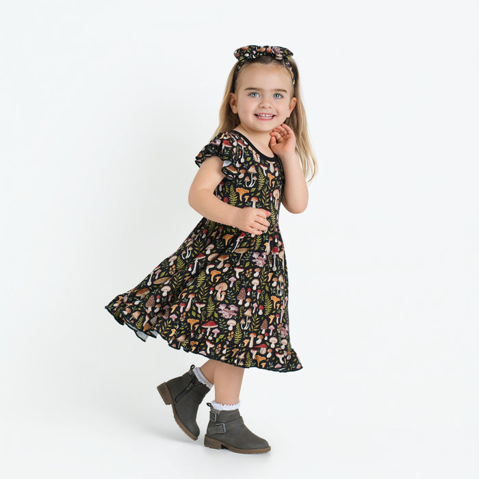 A young girl stands smiling in a Jammie Session Mystic Mushrooms Short Sleeve Big Kid Twirl Dress, paired with a matching headband, gray ankle boots, and white socks against a plain white background.