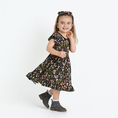 A young girl stands smiling in a Jammie Session Mystic Mushrooms Short Sleeve Big Kid Twirl Dress, paired with a matching headband, gray ankle boots, and white socks against a plain white background.