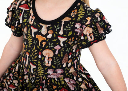 A child wears the Jammie Session Mystic Mushrooms Short Sleeve Big Kid Twirl Dress, featuring soft black fabric with a vibrant mushroom and foliage pattern, shown from shoulders to mid-torso against a white background.