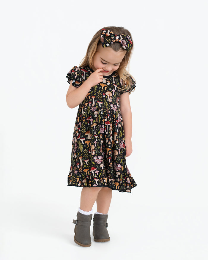 A young child stands against a plain white background wearing the Jammie Session Mystic Mushrooms Short Sleeve Big Kid Twirl Dress, featuring a colorful mushroom print, paired with a matching headband, gray boots, and white socks.