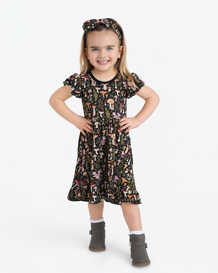 A young child smiles in a Jammie Session Mystic Mushrooms Short Sleeve Big Kid Twirl Dress featuring a colorful mushroom print, paired with a matching headband, grey boots, and white socks against a plain white background.