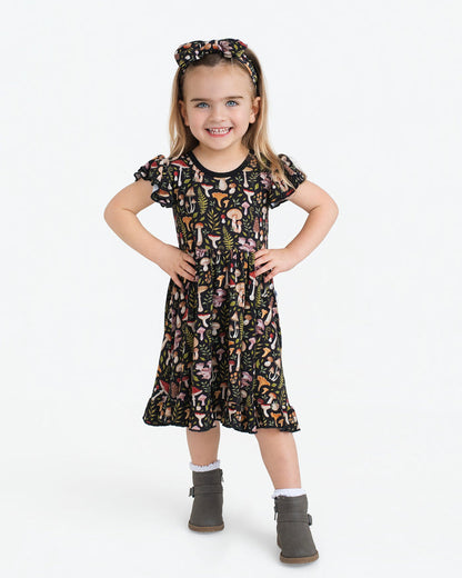 A young child smiles in a Jammie Session Mystic Mushrooms Short Sleeve Big Kid Twirl Dress featuring a colorful mushroom print, paired with a matching headband, grey boots, and white socks against a plain white background.