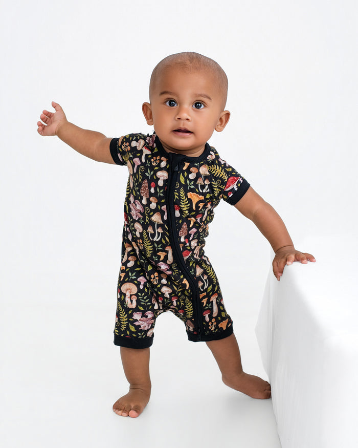 A baby balances with one hand on a white surface, wearing the Jammie Session Mystic Mushrooms Short Sleeve Romper featuring a colorful mushroom and leaf pattern.