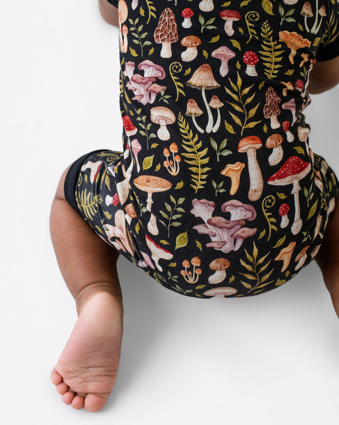 A baby wearing the Jammie Session Mystic Mushrooms Short Sleeve Romper, featuring colorful mushroom and plant patterns, shown from behind on a white background.