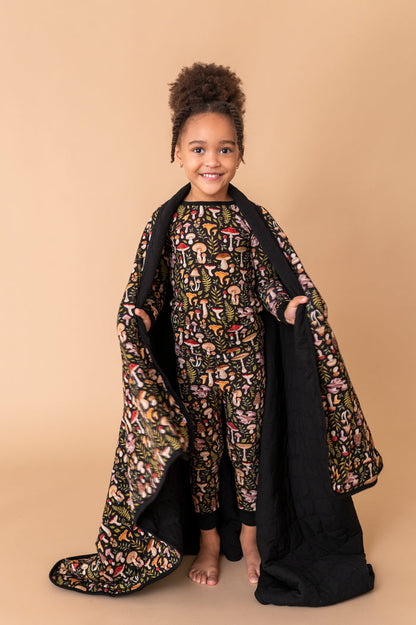 A young child smiles at the camera, standing barefoot on a beige backdrop while holding the Jammie Session Mystic Mushrooms Toddler Quilted Blanket and wearing matching patterned pajamas.