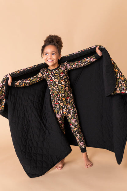 A young child in patterned pajamas stands barefoot, smiling and holding open a Jammie Session Mystic Mushrooms Toddler Quilted Blanket against a beige background.