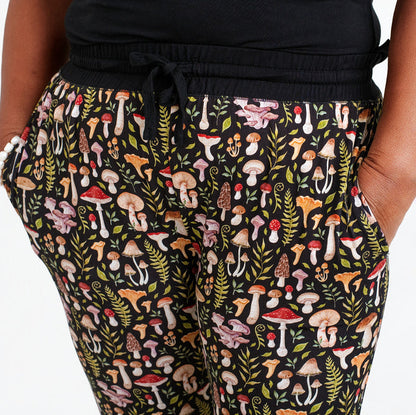 A person wearing Jammie Session's Mystic Mushrooms Women's Jogger Pants, featuring black fabric with vibrant mushroom and leaf patterns, stands with hands in pockets.