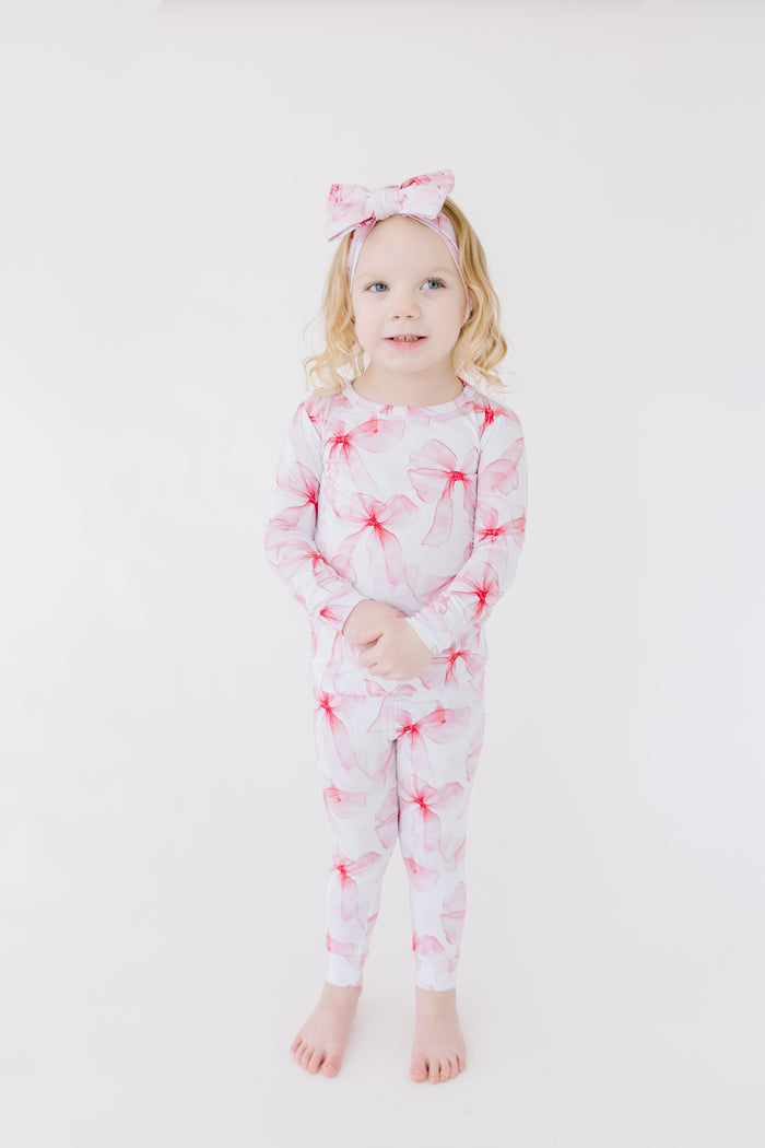 A young child stands barefoot against a white background, wearing Jammie Session's Pretty in Bows Long Sleeve PJ's‚Äîfloral bamboo pajamas with a matching bow headband, gentle on skin and perfect for sensitive sleepers.