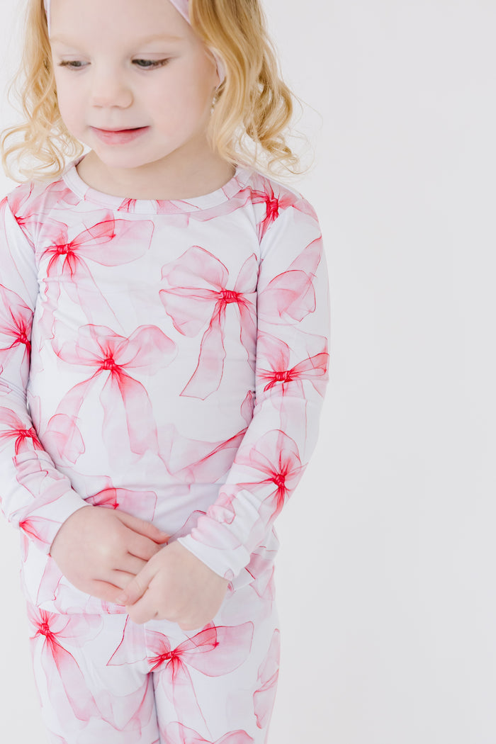 A young blonde child wears Jammie Session‚Äôs Pretty in Bows Long Sleeve PJ's, featuring red bow patterns, standing against a plain white background.