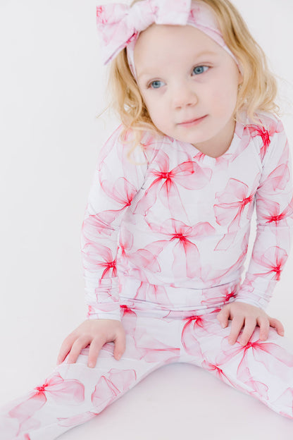 A young child in Jammie Session's Pretty in Bows Long Sleeve PJ's‚Äîa white and pink floral set with a matching headband‚Äîsits on the floor against a plain white background.