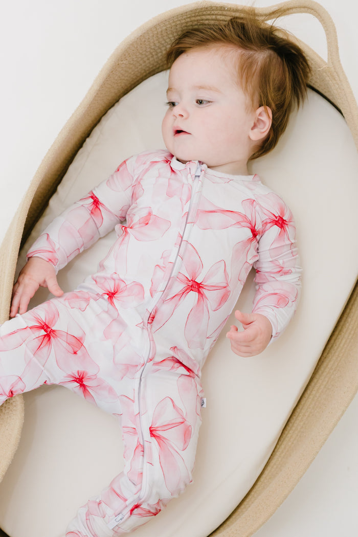 A baby cozies up in a basket, wearing the Jammie Session Pretty in Bows Romper‚Äîa white and pink floral-patterned long sleeve outfit‚Äîlooking off to the side.