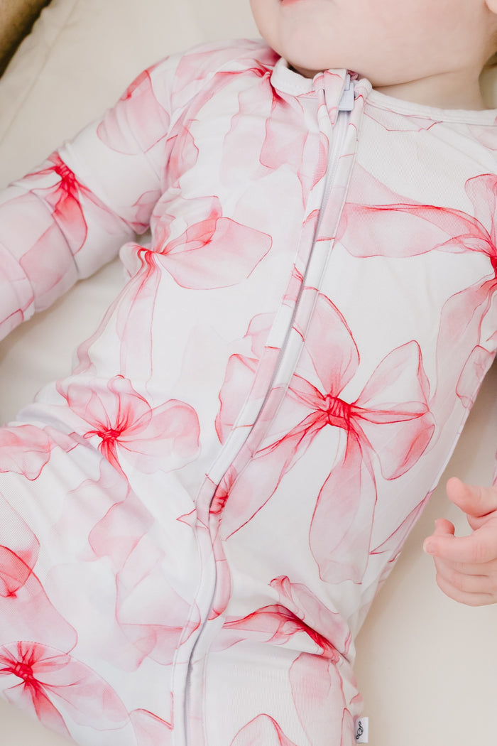 A baby in the Jammie Session Pretty in Bows Romper‚Äîa white bamboo outfit with pink floral patterns‚Äîlies down with one hand partially visible.