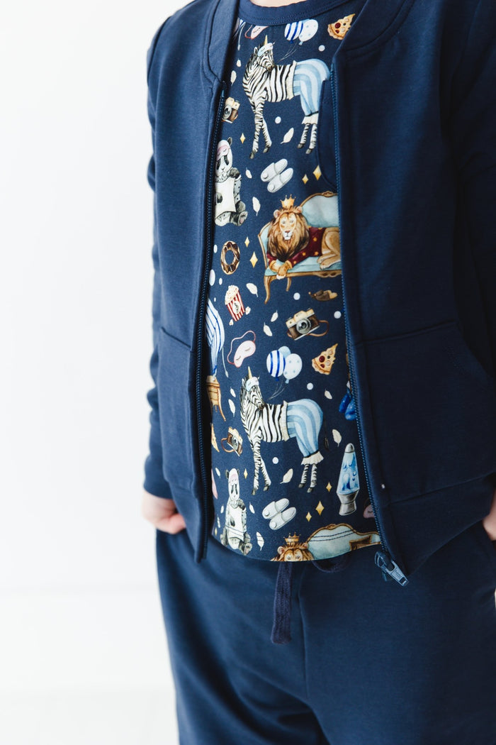 A person wearing the Jammie Session Up Zoo Late Solid Kids' Jacket (runs small, please size up), navy pants with hands in pockets, and an animal-themed shirt with zebras, lions, dogs, and space motifs.
