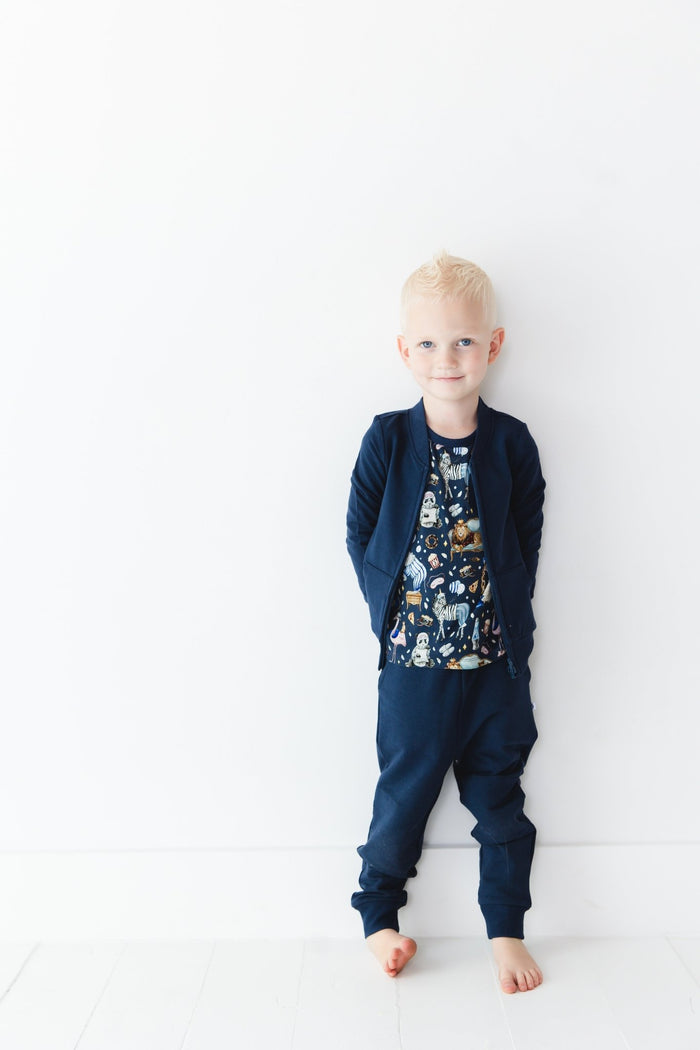 A young blond boy stands barefoot against a white wall, hands in pockets, wearing the Jammie Session Up Zoo Late Solid Kids' Jacket (runs small, please size up) over a graphic t-shirt and looks at the camera.