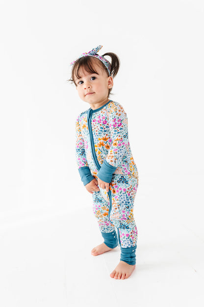 A toddler wearing the Jammie Session Whispering Wildflowers Romper with matching headband stands barefoot on a white background.