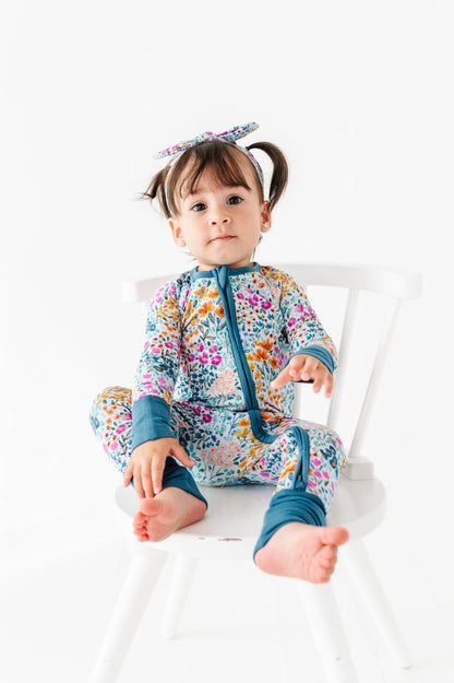 A young child with pigtails sits barefoot on a white chair, wearing the Jammie Session Whispering Wildflowers Romper and a matching headband, set against a plain white background.