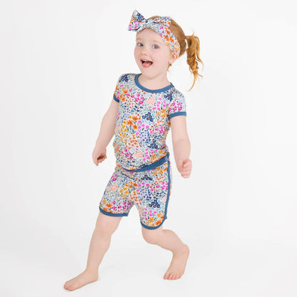 A young red-haired child wears the Jammie Session Whispering Wildflowers Short Pajama Set, featuring a floral-patterned shirt, shorts, and matching headband made from buttery soft bamboo viscose, against a plain white background.