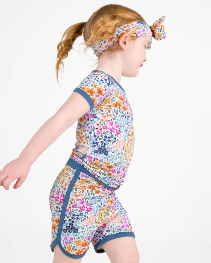 A young child wears the Jammie Session Whispering Wildflowers Short Pajama Set‚Äîfeaturing a colorful floral print, matching headband, and made from buttery soft, hypoallergenic bamboo viscose‚Äîwalking against a plain white background.