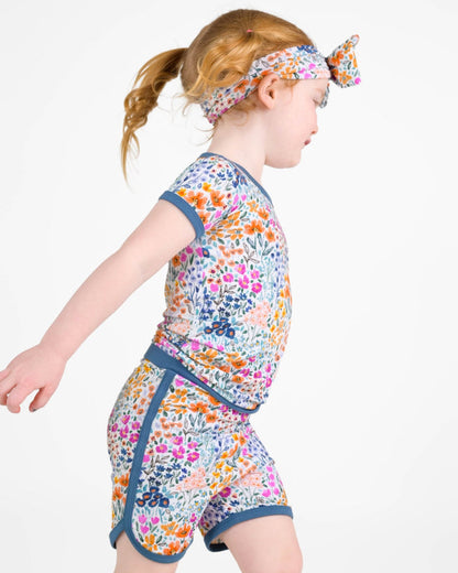 A young child wears the Jammie Session Whispering Wildflowers Short Pajama Set‚Äîfeaturing a colorful floral print, matching headband, and made from buttery soft, hypoallergenic bamboo viscose‚Äîwalking against a plain white background.