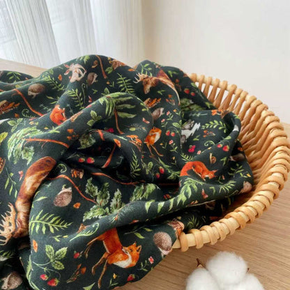 A wicker basket holds the Jammie Session "Wood You Be My Friend" breathable and cozy bamboo cotton muslin swaddle blanket, featuring woodland animal and foliage patterns, placed on a wooden surface next to cotton.