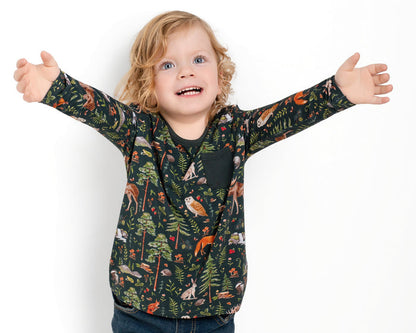 A young child with blonde curly hair wears the Jammie Session "Wood You Be My Friend" Kids' Long Sleeve T-shirt featuring forest animal prints, standing with arms open wide against a plain white background.