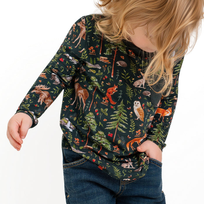 A young child with long blonde hair wearing jeans and a dark "Wood You Be My Friend" Kids' Long Sleeve T-shirt by Jammie Session, featuring a woodland animal and forest print, looks downward.