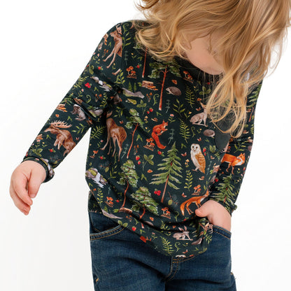 A young child with long blonde hair wearing jeans and a dark "Wood You Be My Friend" Kids' Long Sleeve T-shirt by Jammie Session, featuring a woodland animal and forest print, looks downward.