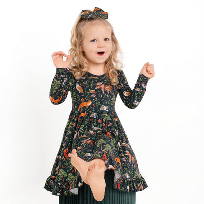 A young girl with blonde hair wears the Jammie Session "Wood You Be My Friend" Long Sleeve Big Kid Twirl Dress and matching bow, standing on one foot with arms raised against a plain white background.