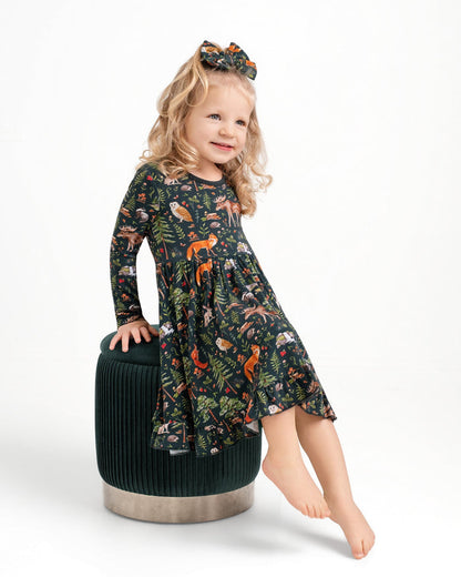 A young blonde girl with a matching bow sits on a green ottoman, wearing the Jammie Session "Wood You Be My Friend" Long Sleeve Big Kid Twirl Dress featuring woodland animals and foliage prints.
